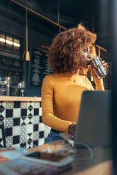 Frau, die an einem Laptop arbeitet, sitzt in einem Restaurant und trinkt Kaffee. Afroamerikanische Frau, die eine Tasse Schokoladenshake genießt, während sie an einem Laptop in einem Café arbeitet. - JLPSF21867
