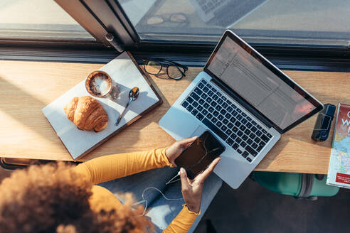 Frau Reisende sitzt in einem Café und arbeitet an einem Laptop und schaut auf ihr Handy. Frau Freiberuflerin überprüft ihr Handy sitzt in einem Restaurant mit Laptop und Snacks. - JLPSF21868