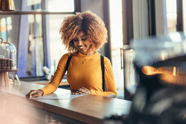 Lächelnde afroamerikanische Frau, die an der Kasse eines Cafés steht und sich die Speisekarte ansieht. Fröhliche afroamerikanische Reisende, die in einem Café Essen bestellt. - JLPSF21869