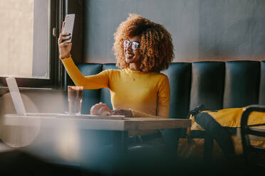 Lächelnde afroamerikanische Frau, die ein Selfie auf dem Handy in einem Restaurant mit Snacks und Laptop auf dem Tisch macht. Weibliche Touristin, die in einem Restaurant speist, während sie am Laptop arbeitet und ein Selfie macht. - JLPSF21878