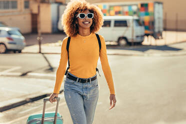 Weibliche afroamerikanische Reisende im Urlaub, die mit ihrem Trolley durch die Stadt läuft. Lächelnde Touristin mit Sonnenbrille und Rucksack, die mit Gepäck auf der Straße läuft. - JLPSF21881