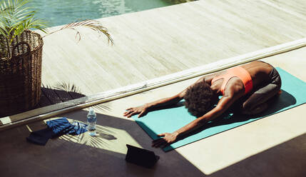 Afrikanische Frau macht Yoga im Freien an einem Swimmingpool. Fitness-Frau übt Kinderpose Yoga auf Übungsmatte am Pool mit einem digitalen Tablet vor. - JLPSF21889