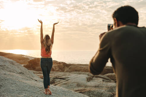 Mann fotografiert mit einem Mobiltelefon eine Frau, die auf einem Felsen am Strand steht. Rückansicht der Frau mit erhobenen Armen und Blick nach oben mit Sonne und Meer im Hintergrund. - JLPSF21900