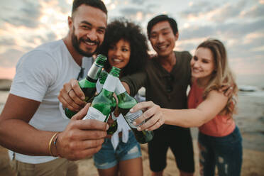 Eine Gruppe multiethnischer Freunde, die zusammen am Strand stehen und Wein genießen. Zwei fröhliche Paare, die am Strand stehen und mit Bierflaschen anstoßen. - JLPSF21903