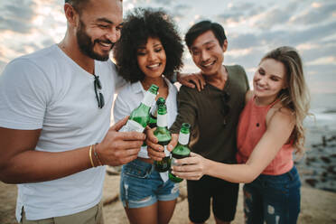 Multiethnische Freunde stehen im Urlaub am Strand und genießen Bier. Vier fröhliche Freunde stehen zusammen am Strand, stoßen mit Bierflaschen an und haben Spaß. - JLPSF21904