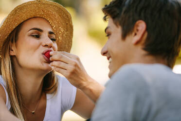 Frau mit Hut, die mit ihrem Freund im Freien sitzt und Erdbeeren isst. Nahaufnahme eines Paares, das zusammen sitzt und Spaß beim Picknick hat. - JLPSF21905