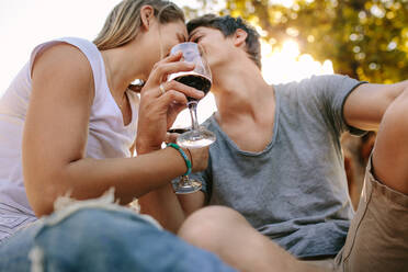 Ein Paar sitzt im Freien und hält ein Glas Wein und küsst sich. Ein Paar sitzt im Park und trinkt Wein unter einem Baum mit Sonnenlicht im Hintergrund. - JLPSF21907