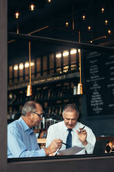 Vertikale Aufnahme von zwei älteren Männern, die sich in einer Kneipe treffen und über ein Dokument diskutieren. Geschäftsleute, die in einem Café am Fenster sitzen und sich mit einem Bericht in der Hand unterhalten. - JLPSF21913