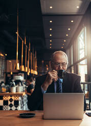 Vertikale Aufnahme eines älteren Geschäftsmannes, der auf einen Laptop schaut und einen Kaffee im Café trinkt. Älterer Mann im Geschäftsanzug, der mit einem Laptop am Cafétisch sitzt und Kaffee trinkt. - JLPSF21917