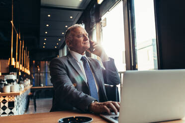 Senior businessman at coffee shop making a phone call. Mature businessman talking on mobile phone from a modern coffee shop with laptop on table. - JLPSF21919