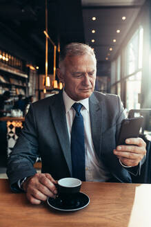Älterer Geschäftsmann, der eine Textnachricht auf seinem Telefon liest, während er im Café sitzt und Kaffee trinkt. Reifer Unternehmer, der in einem Café eine Textnachricht auf seinem Smartphone schreibt. - JLPSF21920