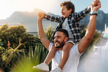 Fröhlicher Mann, der seinen Sohn auf den Schultern trägt und Spaß hat. Vater und Sohn im Urlaub, die im Freien spazieren gehen, mit Sonne im Hintergrund. - JLPSF21925