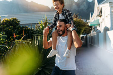 Glücklicher Mann mit seinem Sohn auf den Schultern, der auf der Straße spazieren geht. Vater und Sohn in fröhlicher Stimmung im Urlaub, die im Freien mit der Sonne im Hintergrund spazieren gehen. - JLPSF21926