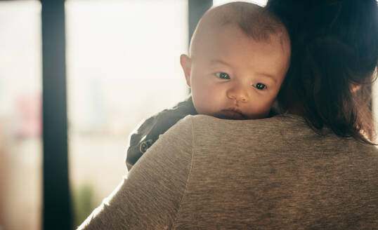 Rückansicht einer Mutter, die ihr Baby trägt. Das Baby bleibt ruhig und stützt sein Kinn auf die Schulter seiner Mutter. - JLPSF21932