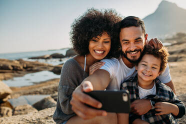 Lächelndes Paar mit Kind, das auf einem Felsen in der Nähe der Küste sitzt und für ein Selfie posiert; Mann, der ein Selfie mit seiner Familie macht, indem er ein Handy benutzt und im Freien sitzt. - JLPSF21933
