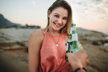 Lächelnde Frau im Urlaub am Strand mit einer Weinflasche in der Hand. Fröhliche Frau, die mit einem Freund am Meer stehend auf eine Flasche Bier anstößt. - JLPSF21938