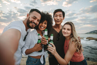 Cheerful friends on vacation at the beach taking a selfie holding bottles of beer. Two couples standing at the beach in cheerful mood enjoying a holiday. - JLPSF21939