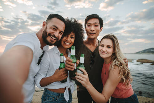 Fröhliche Freunde im Urlaub am Strand machen ein Selfie mit Bierflaschen in der Hand. Zwei Paare, die am Strand stehen und in fröhlicher Stimmung ihren Urlaub genießen. - JLPSF21939