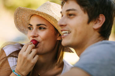 Lächelndes Paar, das im Freien zusammensitzt und wegschaut. Frau mit Hut isst eine Erdbeere und sitzt mit ihrem Freund in einem Park. - JLPSF21946