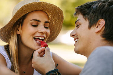 Mann füttert seine Freundin mit einer Erdbeere im Freien sitzend. Frau mit Hut isst die von ihrem Freund gefütterte Erdbeere. - JLPSF21948