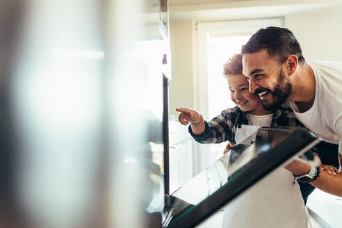 Ein lächelnder Vater und sein Sohn betrachten das Essen in einem Ofen. Ein Mann öffnet die Tür eines Ofens und überprüft das Essen mit seinem Kind. - JLPSF21955