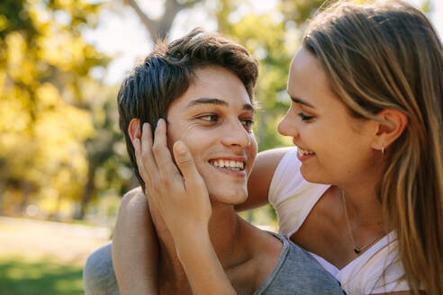 Glückliches Paar im Park, das sich lächelnd ansieht. Der Mann trägt seine Freundin auf dem Rücken, während die Frau sein Gesicht liebevoll berührt. - JLPSF21956