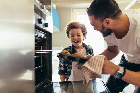 Ein Mann zeigt seinem Sohn gebackene Kekse, während er sie aus dem Ofen nimmt. Ein Kind betrachtet die gebackenen Kekse, die aus dem Ofen kommen, aufgeregt. - JLPSF21960