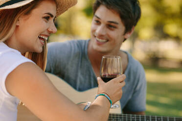 Lächelnde Frau mit Hut, die mit ihrem Freund zusammensitzt und ein Glas Wein in der Hand hält; Mann spielt Gitarre und sitzt mit einer Frau im Park. - JLPSF21968