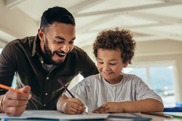 Glücklicher Vater und Sohn sitzen am Tisch und haben Spaß beim Zeichnen mit Buntstiften. Das Kind macht eine Zeichnung in einem Buch, während sein Vater ihn anfeuert. - JLPSF21973