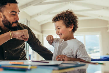 Smiling kid giving a fist bump to his father sitting at a table. Happy father and son sitting at home having fun drawing. - JLPSF21974