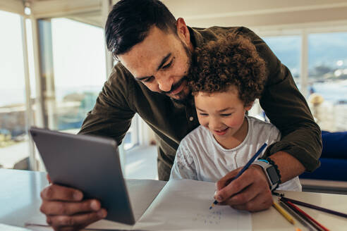 Vater unterrichtet sein Kind, das einen Tablet-PC hält, während es in ein Buch schreibt. Nahaufnahme eines Mannes, der in ein Buch schreibt, während ein lächelndes Kind es ansieht. - JLPSF21977