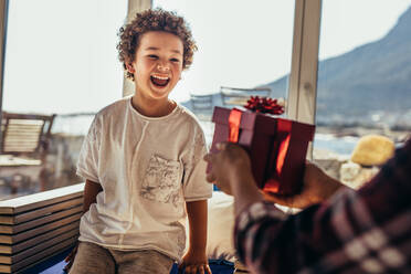 Person giving a gift box to a boy sitting in a beach house. Excited kid shouting in joy seeing a birthday gift being given to him. - JLPSF21989