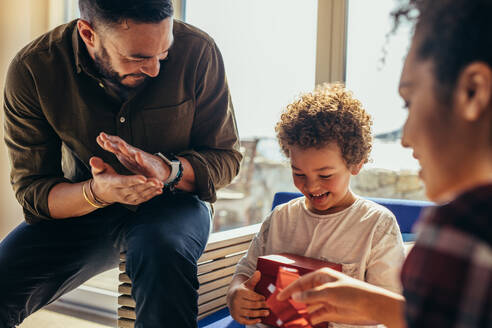Lächelnder Junge, der sein Geburtstagsgeschenk betrachtet und mit seinen Eltern zusammensitzt. Mann und Frau sitzen mit ihrem Kind und feiern seinen Geburtstag in einem Strandhaus. - JLPSF21990