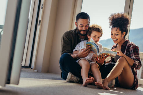 Lächelnder Mann mit Frau und Kind, die Zeit miteinander verbringen und ein Buch lesen. Glückliches Paar sitzt mit seinem Kind auf dem Boden und liest eine Geschichte. - JLPSF21992