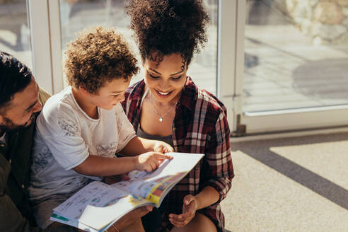 Ein Junge sitzt auf dem Schoß seiner Eltern und hat Spaß beim Lesen eines Märchenbuchs. Ein lächelndes Paar sitzt mit seinem Kind zu Hause und liest ein Märchenbuch. - JLPSF21993