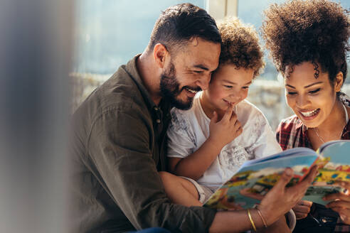 Glückliche Familie, die sich zu Hause beim Lesen eines Märchenbuchs amüsiert. Ein Junge sitzt mit seinem Vater und seiner Mutter und genießt ein Märchenbuch. - JLPSF21994