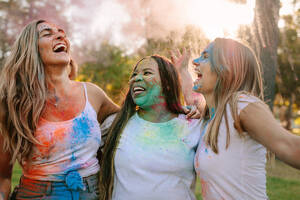 Drei Frauen amüsieren sich beim Holi-Spielen mit Farbklecksen im Gesicht. Freunde genießen Holi in einem Park und lachen vor Freude. - JLPSF21999