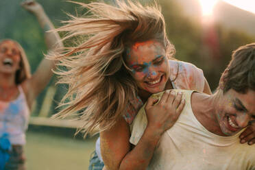 Smiling woman having fun with her boyfriend while playing holi in a park. Woman holding her man from behind while playing holi with friends cheering in the background. - JLPSF22003