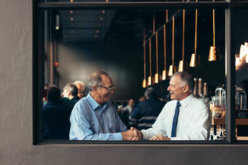 Geschäftsleute sitzen im Café und schütteln sich die Hände. Zwei reife Geschäftsmänner schütteln sich die Hände, während sie in einem Restaurant sitzen. - JLPSF22015