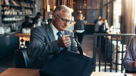 Ein älterer Geschäftsmann sitzt an einem Cafétisch und holt seinen Laptop aus der Tasche, während ein Mann daneben sitzt. - JLPSF22021