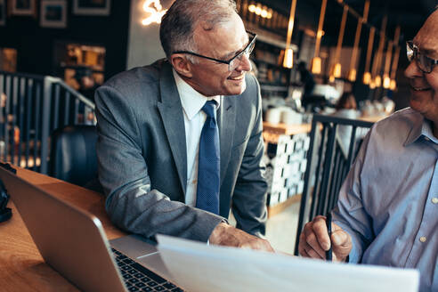 Älterer Geschäftsmann im Gespräch mit männlichem Partner in einem modernen Café. Zwei ältere Geschäftsleute arbeiten zusammen an einem Geschäftsbericht. - JLPSF22024