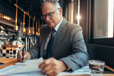 Älterer Geschäftsmann sitzt an einem Cafétisch, liest ein Dokument und macht sich Notizen. Älterer Mann im Anzug arbeitet an einem neuen Projektbudget. - JLPSF22033