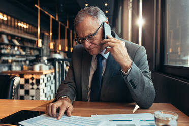 Senior businessmen sitting at cafe talking on mobile phone to discuss business figures. Mature businessmen at coffee shop reading document and making a phone call. - JLPSF22036