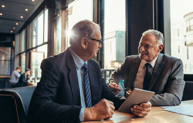 Zwei ältere Geschäftsleute, die ein informelles Treffen in einem Café haben. Ein Geschäftsmann, der ein digitales Tablet hält, spricht mit seinem männlichen Partner, während er an einem modernen Kaffeehaustisch sitzt. - JLPSF22042