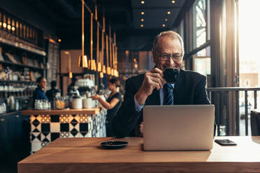 Lächelnder älterer Geschäftsmann, der im Café sitzt, eine Tasse Kaffee trinkt und auf den Laptop schaut. Männlicher Unternehmer im Café mit Laptop. - JLPSF22044