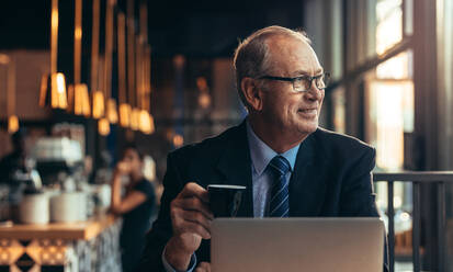 Entspannter älterer Geschäftsmann, der mit einem Kaffee in der Hand in einem Café sitzt und lächelnd wegschaut. Reifer männlicher Unternehmer, der sich in einem Café mit einem Laptop vor sich entspannt. - JLPSF22045