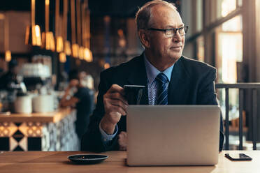 Älterer Geschäftsmann sitzt im Restaurant mit Kaffee und schaut weg. Älterer Unternehmer sitzt im Café mit Laptop auf dem Tisch. - JLPSF22046