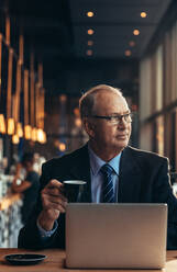 Vertical shot of senior businessman looking away and having coffee. Mature man in sitting at cafe table with laptop drinking coffee. - JLPSF22047