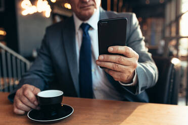 Close up of mobile phone in hand of a businessman with coffee on cafe table. Senior businessman using smartphone at coffee shop. - JLPSF22048