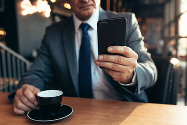 Nahaufnahme eines Mobiltelefons in der Hand eines Geschäftsmannes mit Kaffee auf einem Cafétisch. Älterer Geschäftsmann benutzt Smartphone in einem Café. - JLPSF22048